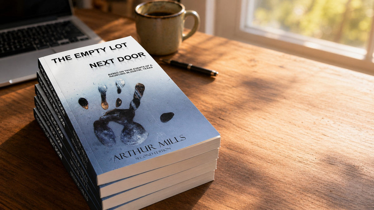 Stack of new paperback copies of The Empty Lot Next Door by Arthur Mills on a sunlit wooden desk near a window.